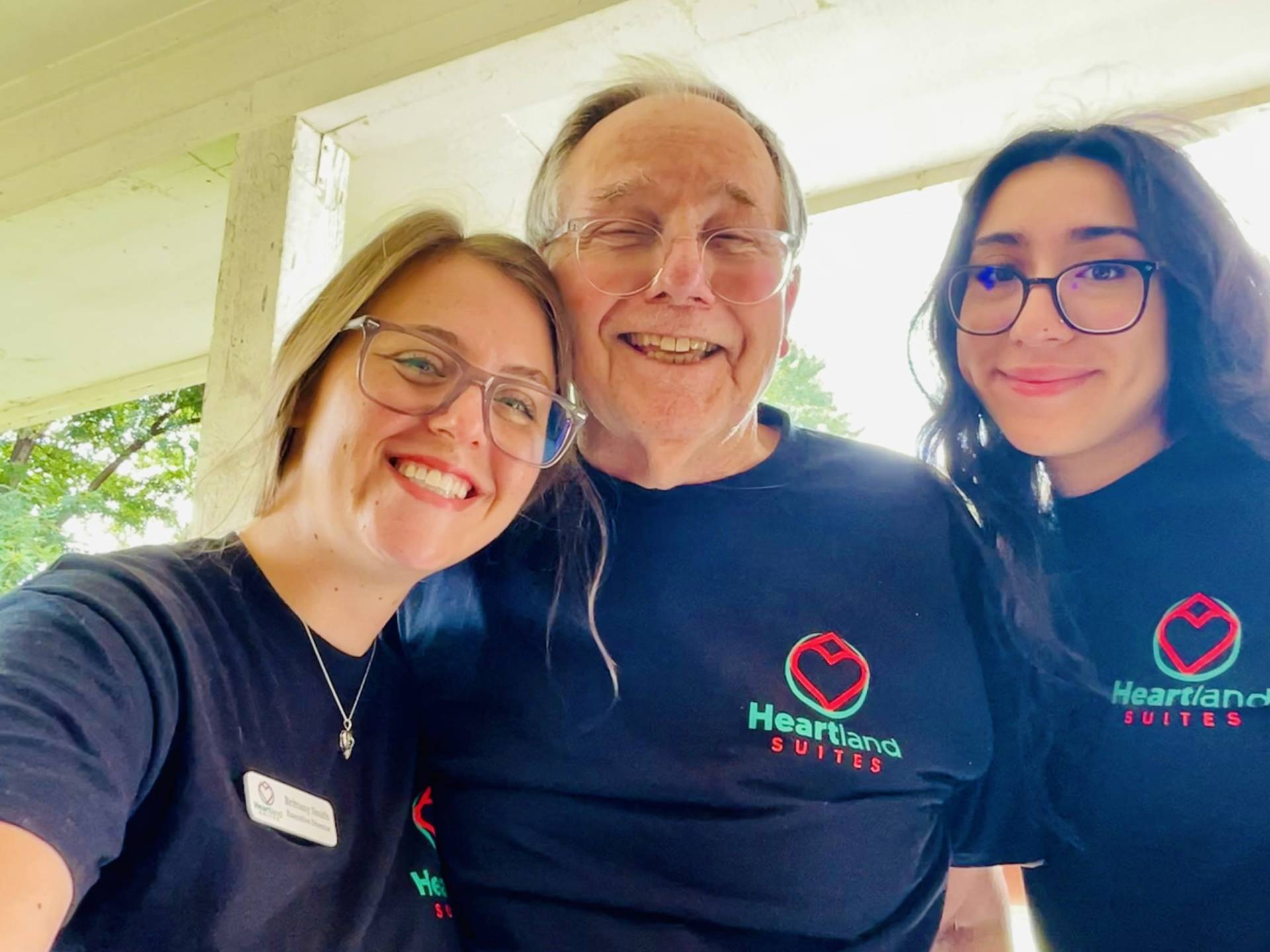 Resident smiling wearing a Heartland Suites shirt with two friendly staff members at a residential assisted living home in Pryor, Oklahoma.